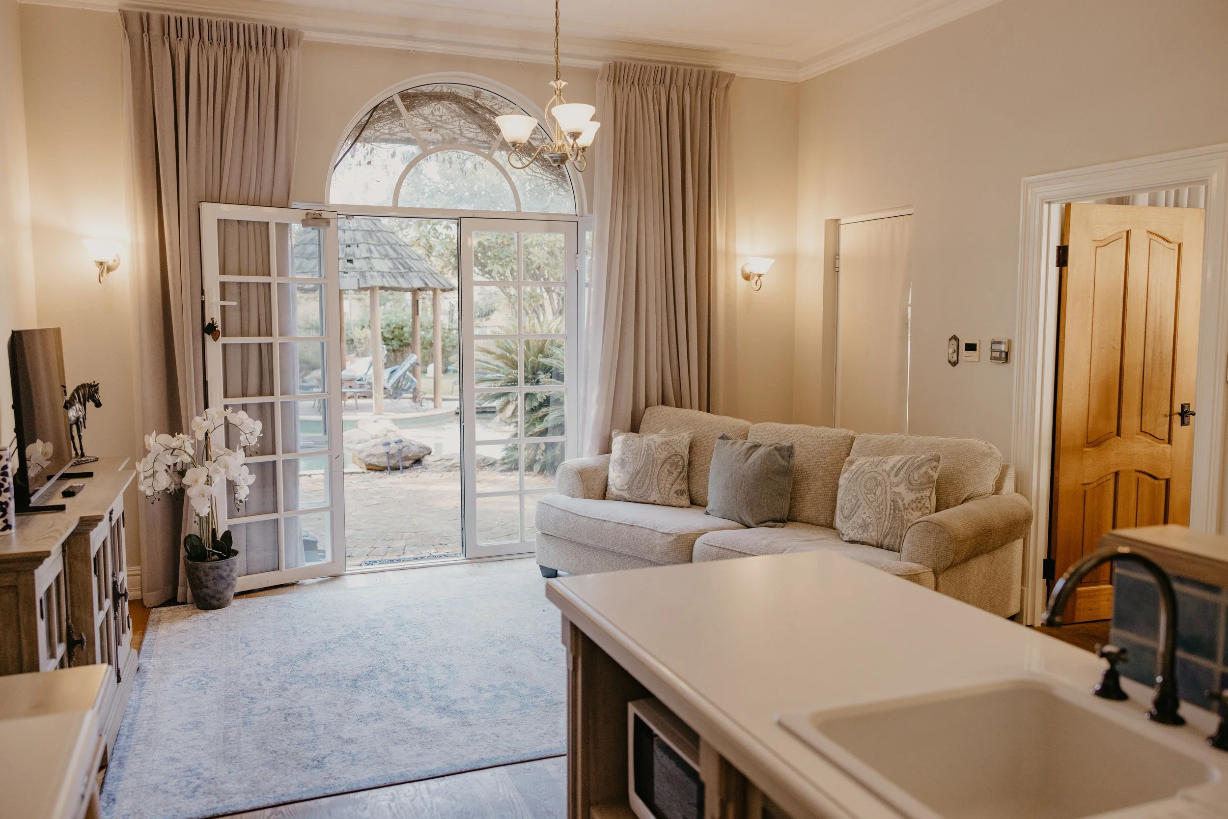 A cozy living room with a large arched window, beige curtains, a chandelier, wall sconces, a beige sofa with patterned pillows, a wooden door, and a view of a backyard with a wooden pavilion and plants.