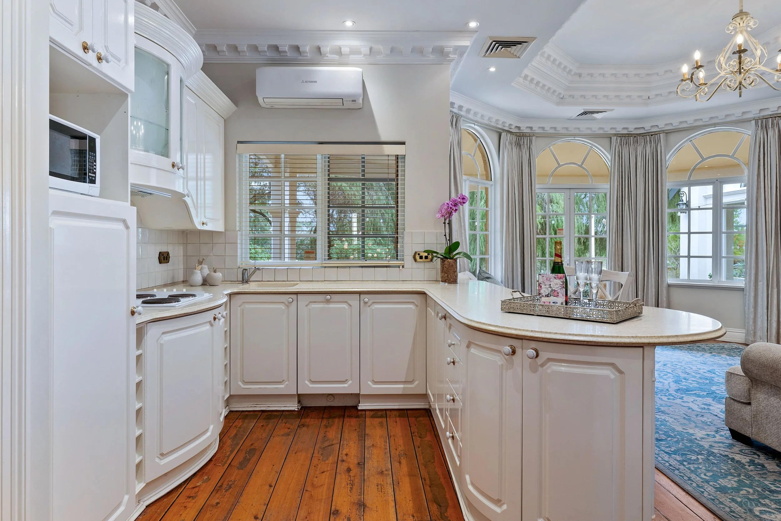 A bright kitchen with white cabinets and a curved kitchen island. Large arched windows with curtains let in natural light. There is a pink orchid plant on the counter and a silver tray with drinkware. The ceiling has crown molding and a chandelier. T