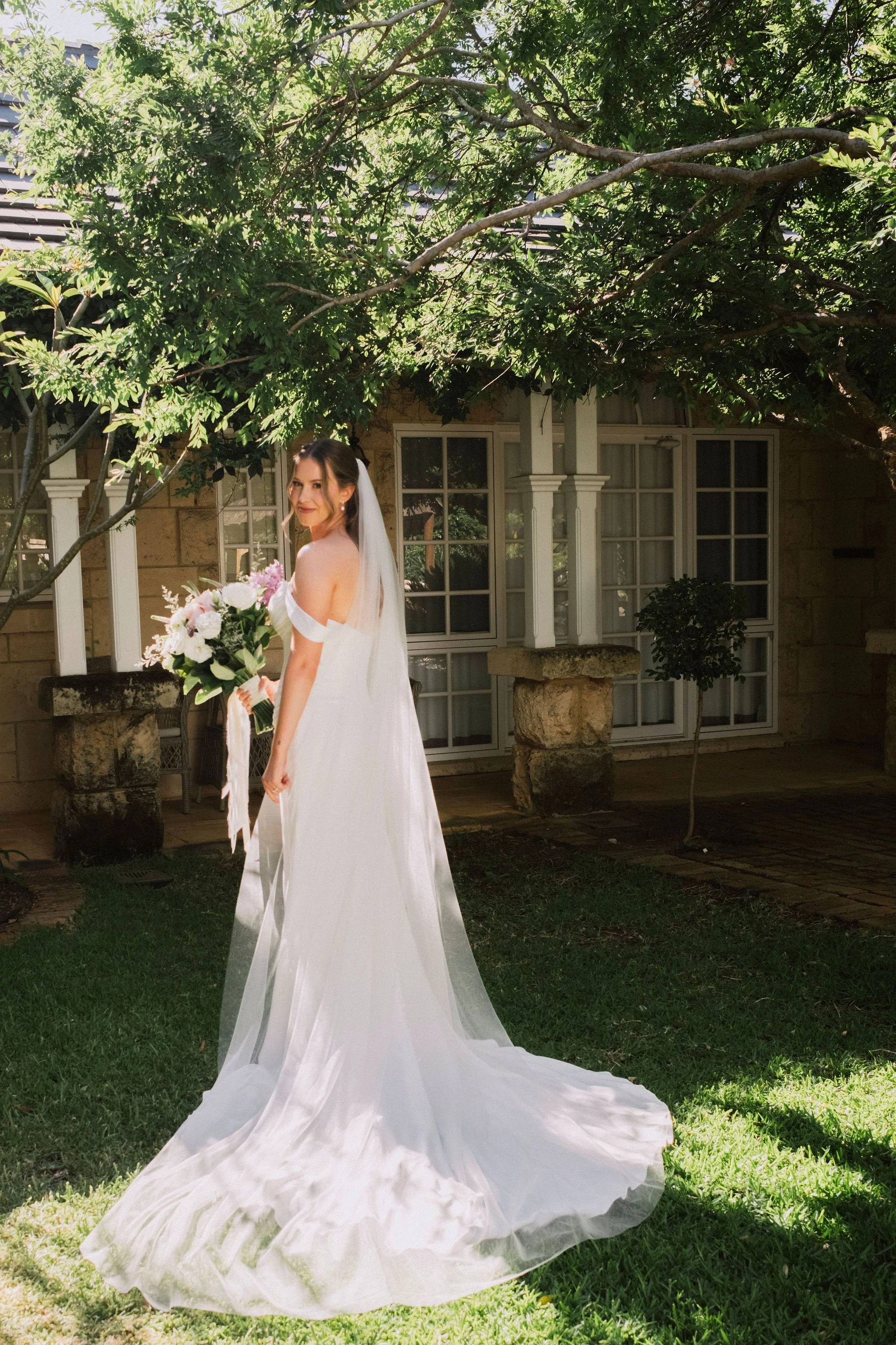 A bride in a white wedding dress holding a bouquet of pink and white flowers, standing on a green lawn under a tree, with a stone and wooden porch in the background.