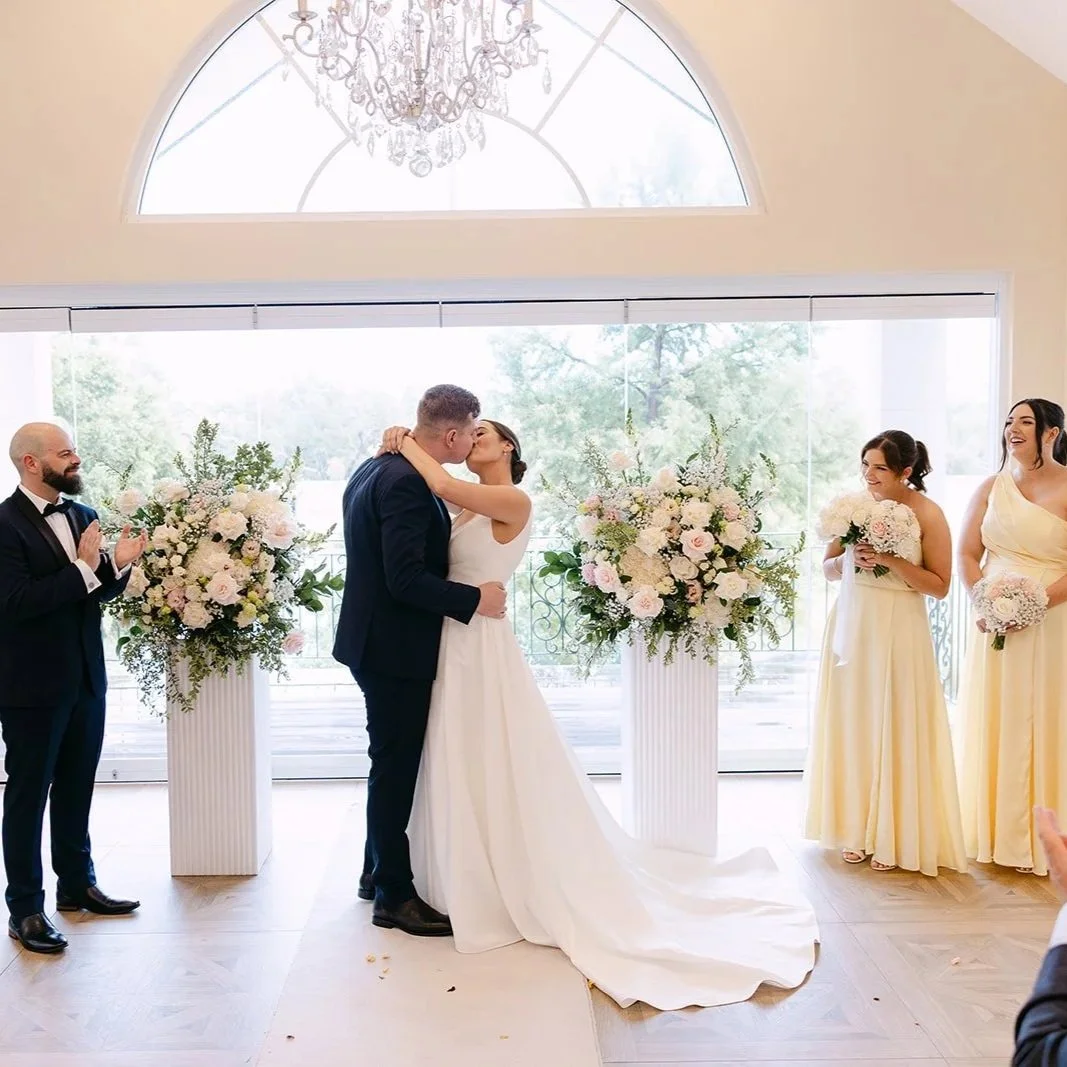 A couple shares a kiss at their wedding ceremony with two bridesmaids and a groomsman standing nearby, decorated with large floral arrangements in an elegant indoor space with large windows and a chandelier.