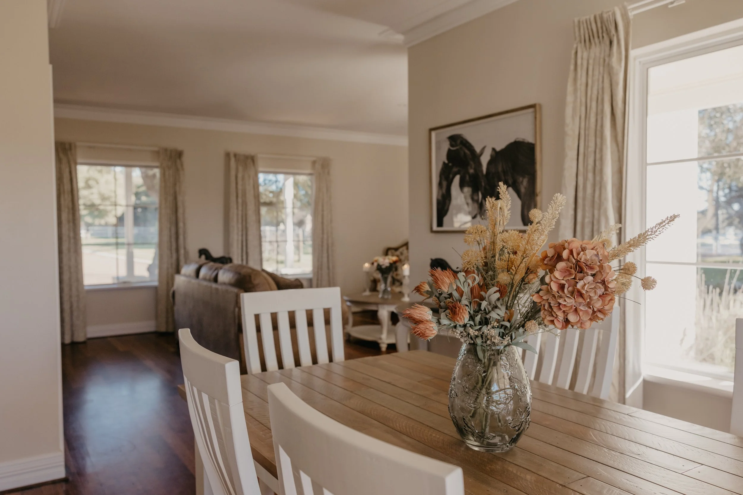 A dining room with a wooden table and white chairs, a glass vase with beige and pink flowers, and large windows with cream curtains letting in natural light.