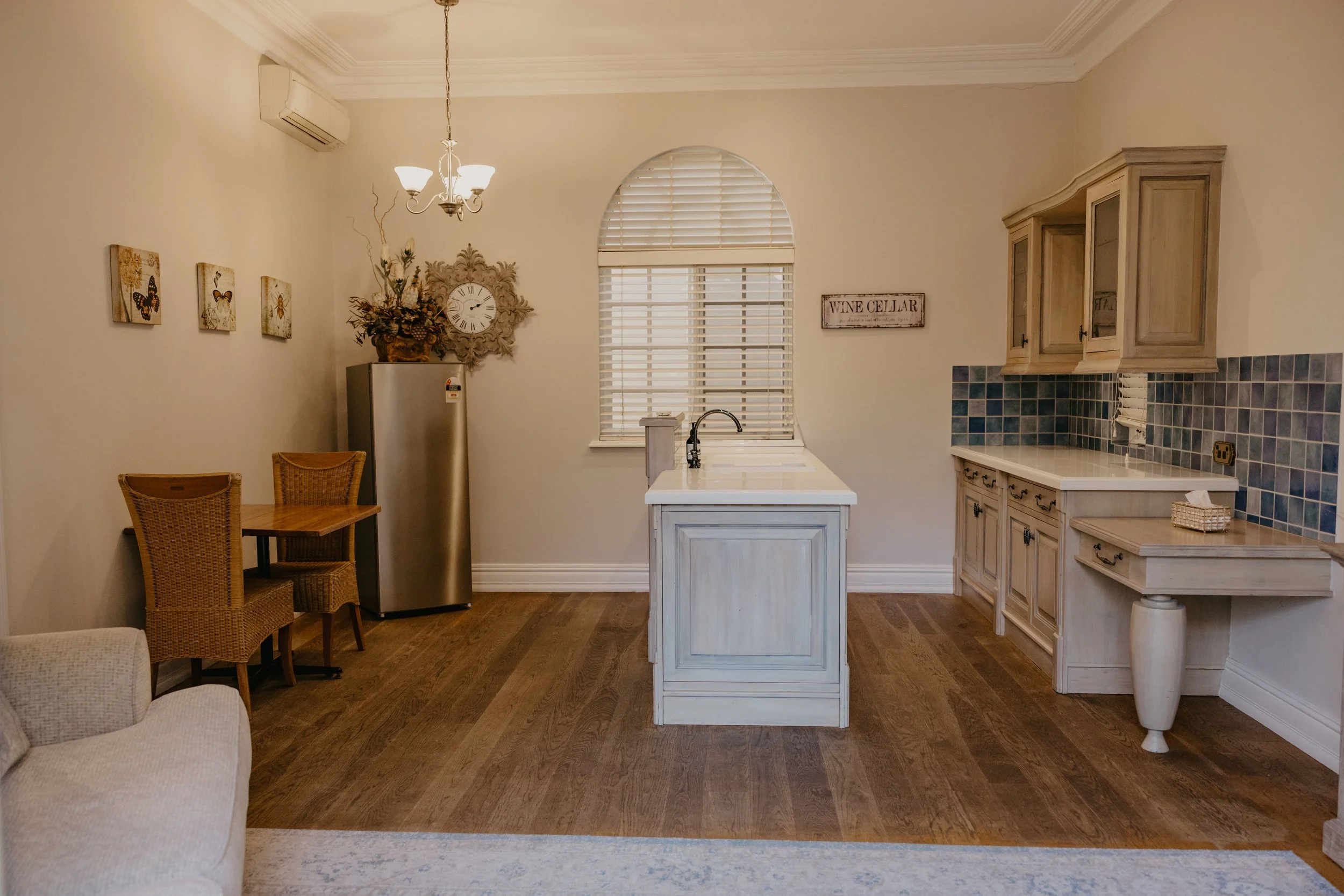 A kitchen and dining area with white cabinets, a chandelier, wall art with butterflies, a small table with two chairs, a stainless steel refrigerator, a window with blinds, and a 