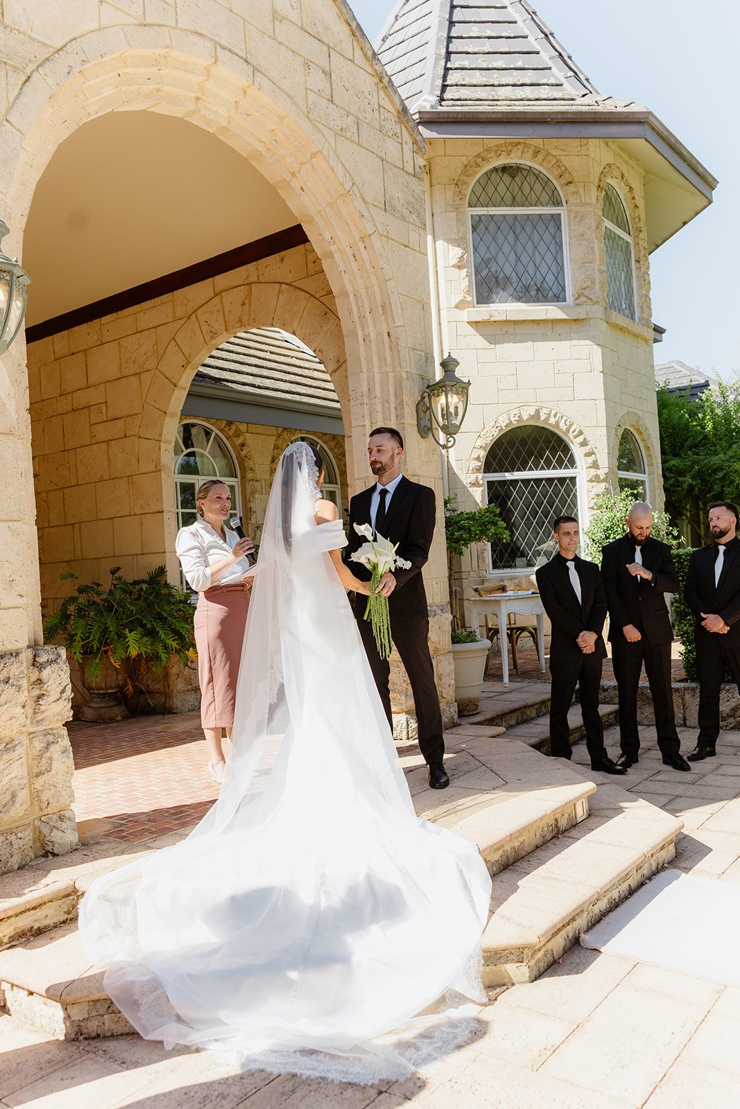 A wedding ceremony taking place outside a large stone house with arched windows and a turret. A bride in a white wedding gown with a long train and veil is holding a bouquet of white lilies while facing a groom in a black suit. A woman officiant hold