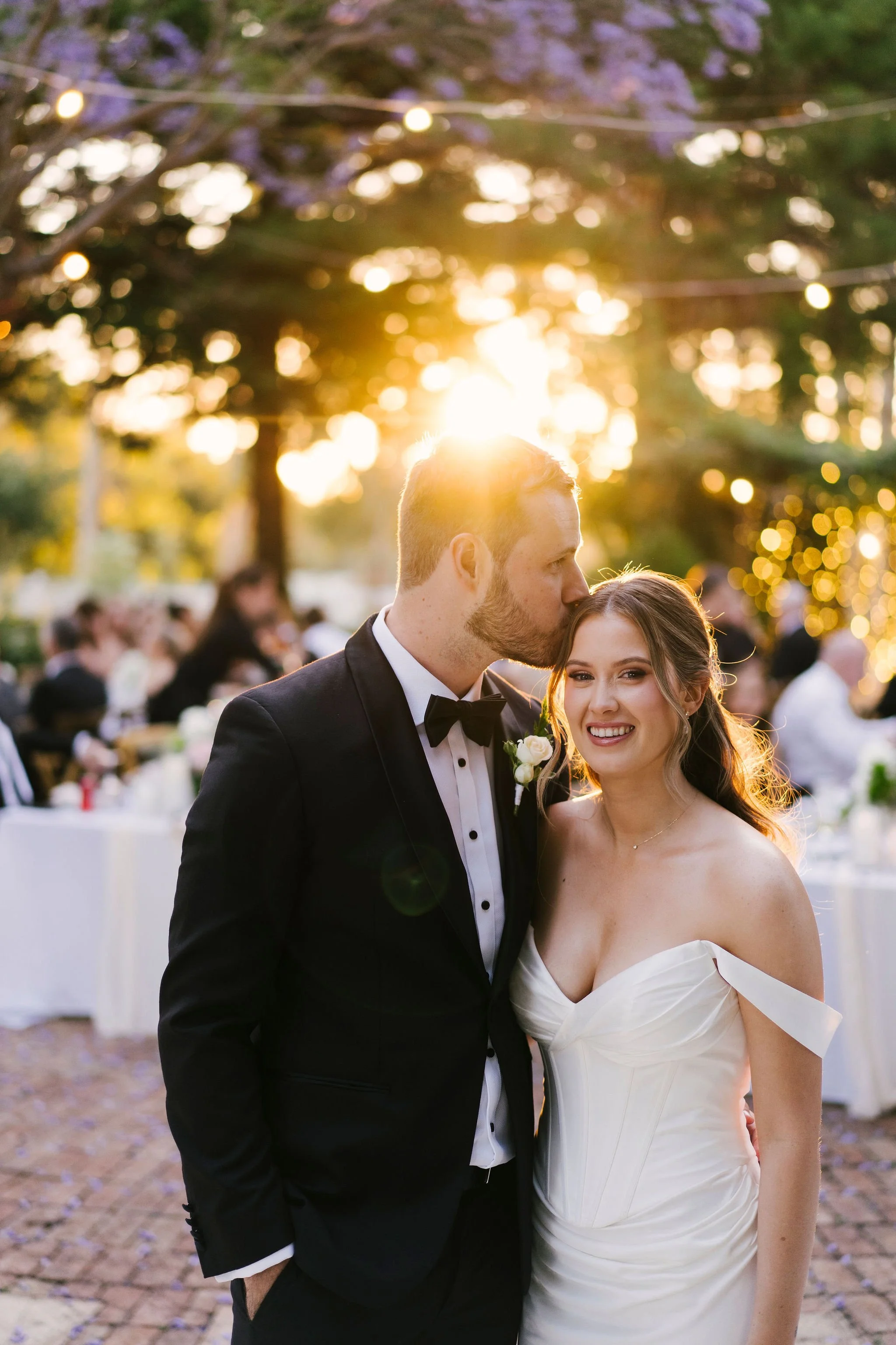 A newlywed couple at sunset, the groom wearing a black tuxedo and the bride in a white wedding dress, standing outdoors with guests in the background.