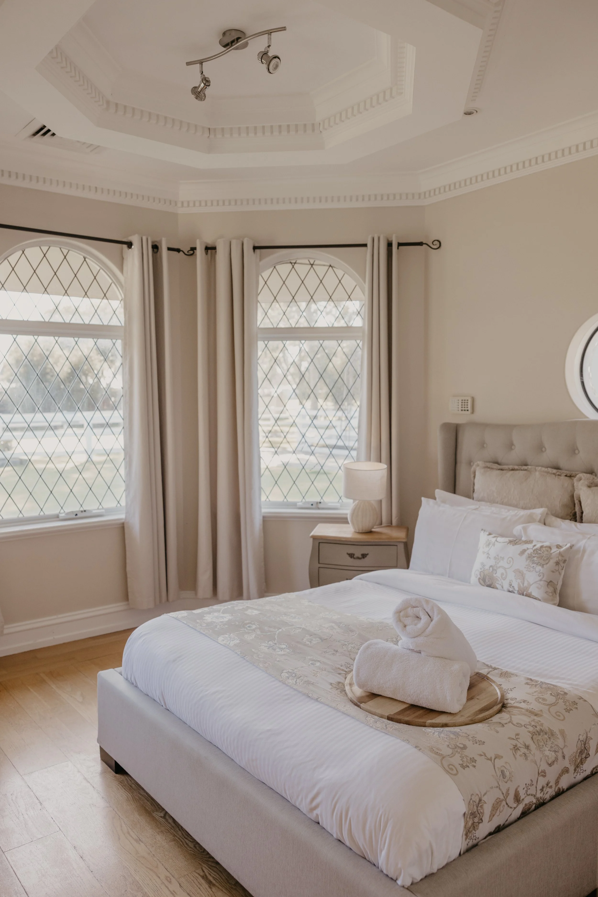 A neatly made bed with a beige and gold embroidered bed runner and matching pillows, a towel folded into the shape of a swan on a wooden tray, beside a nightstand with a beige lamp, large windows with diamond-patterned grillwork and cream curtains, a