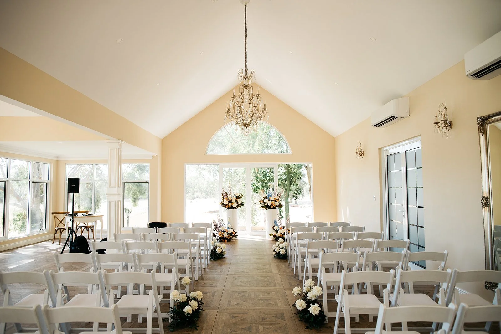 Wedding ceremony setup with white chairs arranged in rows, floral arrangements at the front, large windows, a chandelier hanging from the ceiling, and a bright, airy interior.