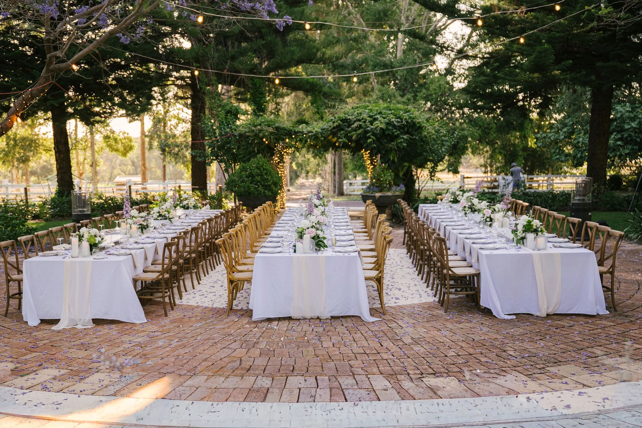 Outdoor wedding reception setup with long white-draped tables, floral centerpieces, surrounded by wooden chairs, string lights hanging from trees, and a brick pathway in a lush garden.