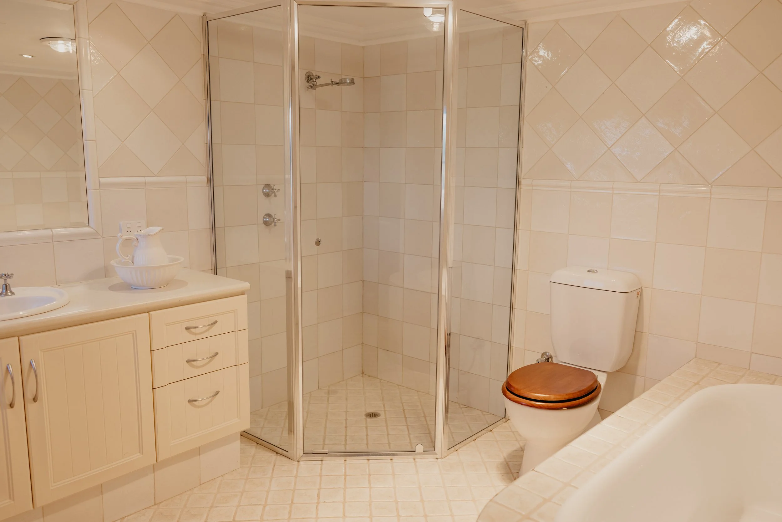 A bathroom with cream-colored tiled walls and floor. It features a corner shower with a glass enclosure, a white vanity with drawers, a mirror, a white sink, a brass faucet, a white bowl, and a toilet with a wooden seat cover.