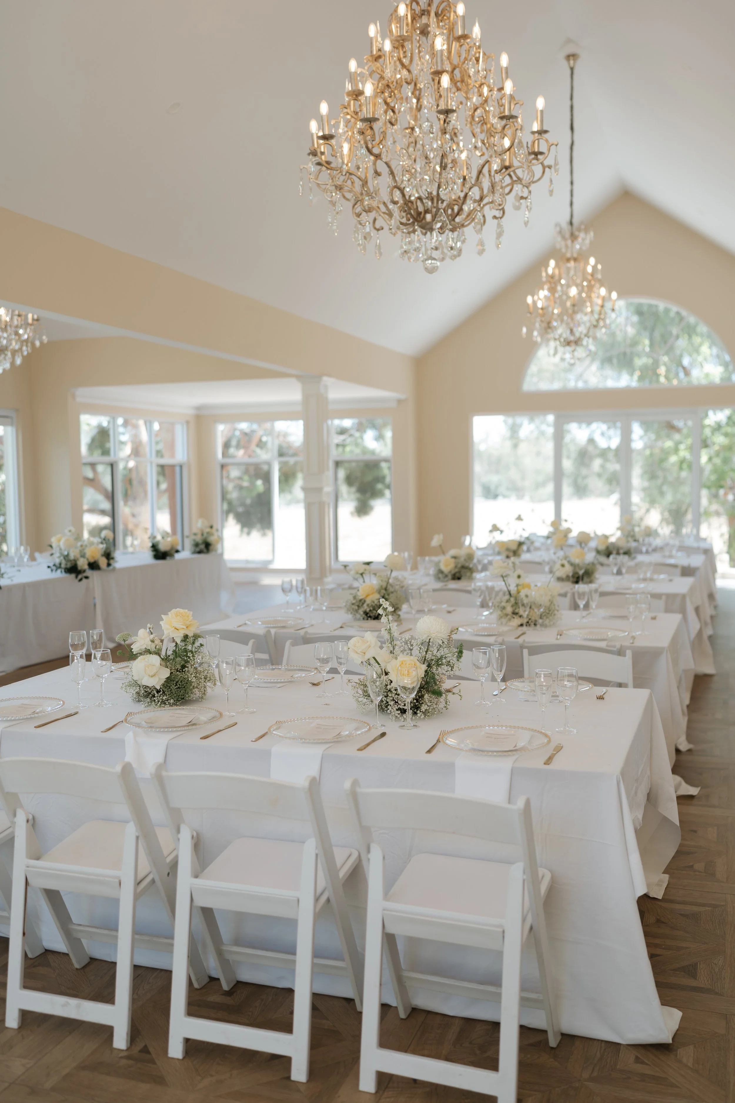 Elegant dining room decorated for a wedding or special event, featuring long tables with white tablecloths, floral centerpieces, and chandeliers hanging from the ceiling.