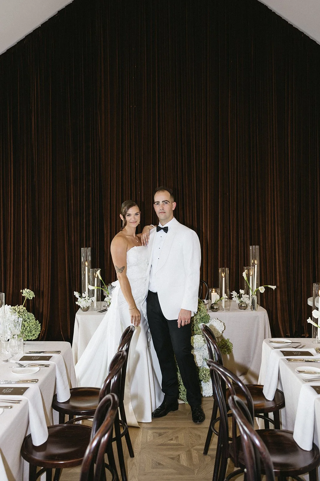 A bride and groom in wedding attire stand together in a decorated reception hall with dark wood paneling background, white tables, elegant flower arrangements, and candles.