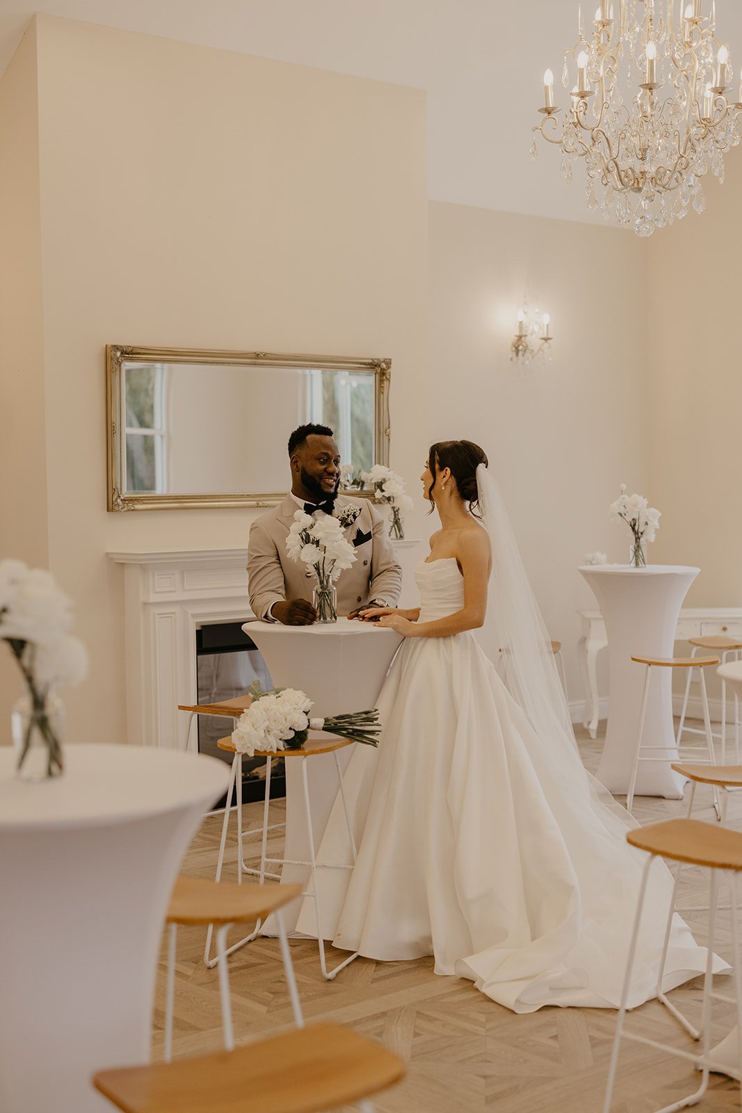 A bride and groom with dark and light skin tones stand at a small white table with white flowers in vases, smiling at each other in a bright, elegant room with white walls, a large mirror, and a chandelier.
