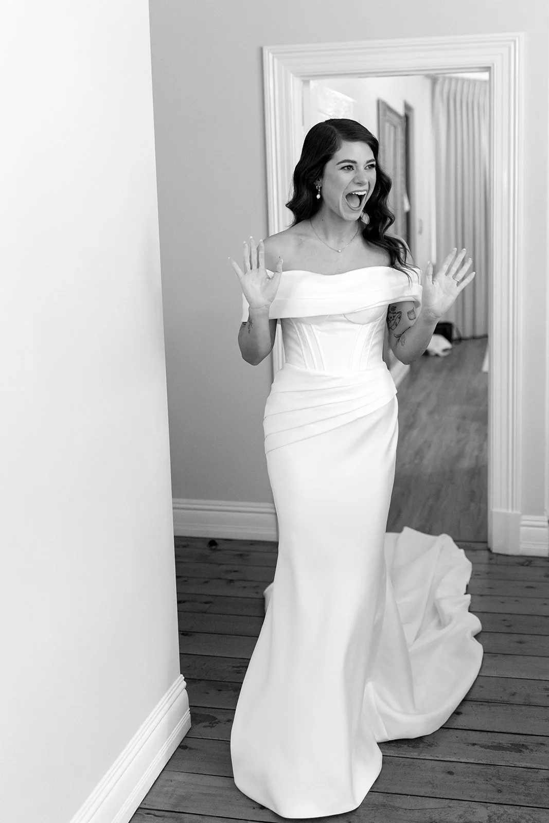 A woman in a white wedding dress standing in front of a mirror with a joyful expression, hands raised, in a home interior with hardwood floors.