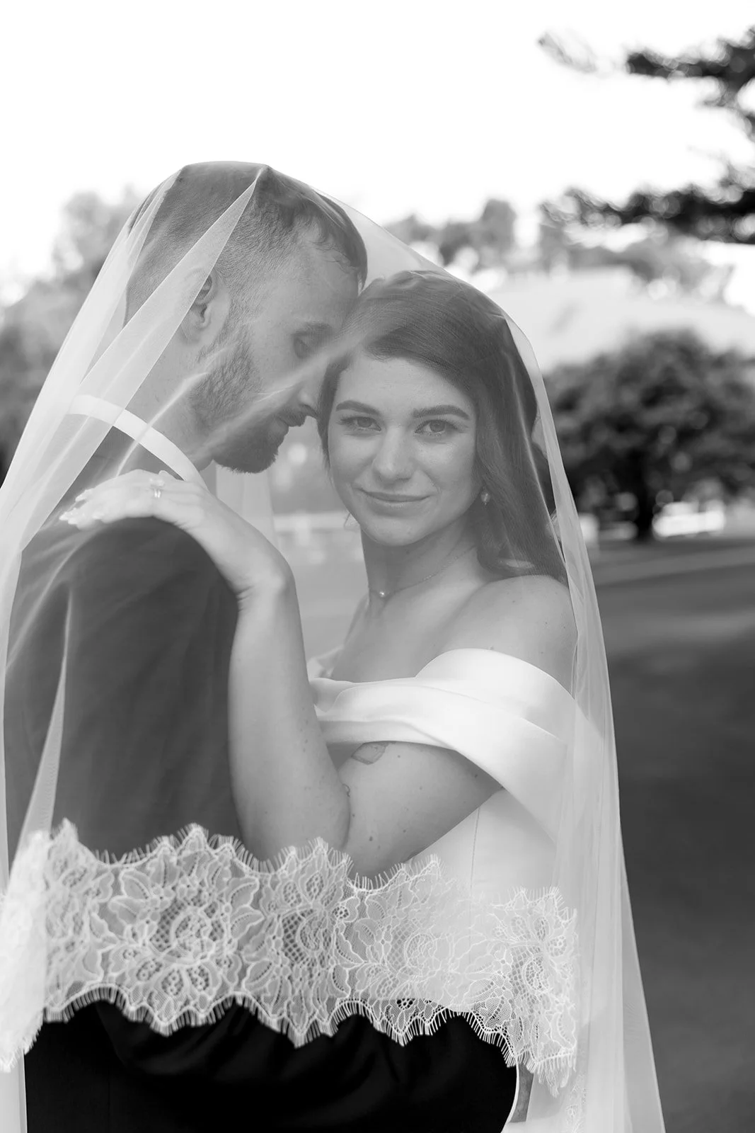 A bride and groom sharing a close moment behind a veil outdoors, with trees in the background.