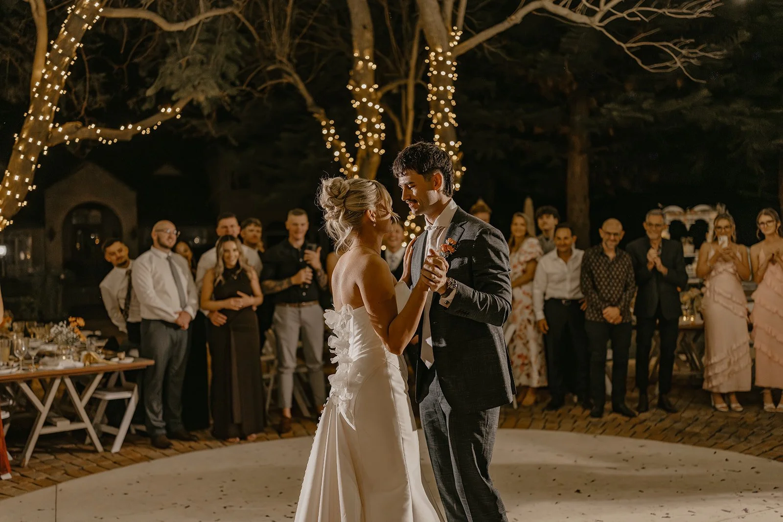 A bride and groom dancing at their wedding reception outdoors at night, surrounded by guests under string lights in a wooded area.