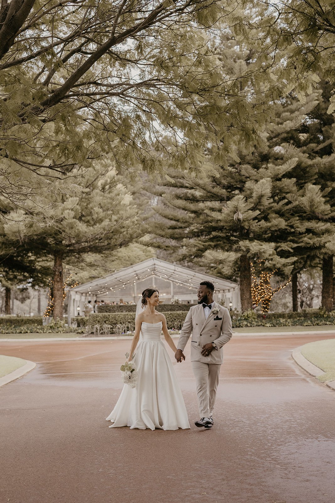 Bride and groom walking hand in hand outdoors, with trees and a decorated tent in the background, at their wedding reception.