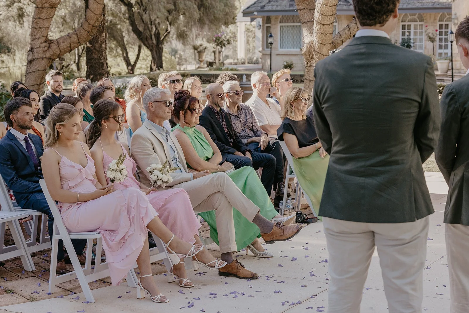 Guests seated outdoors at a wedding ceremony, some holding bouquets, facing a couple standing in front of them, with trees and a building in the background.
