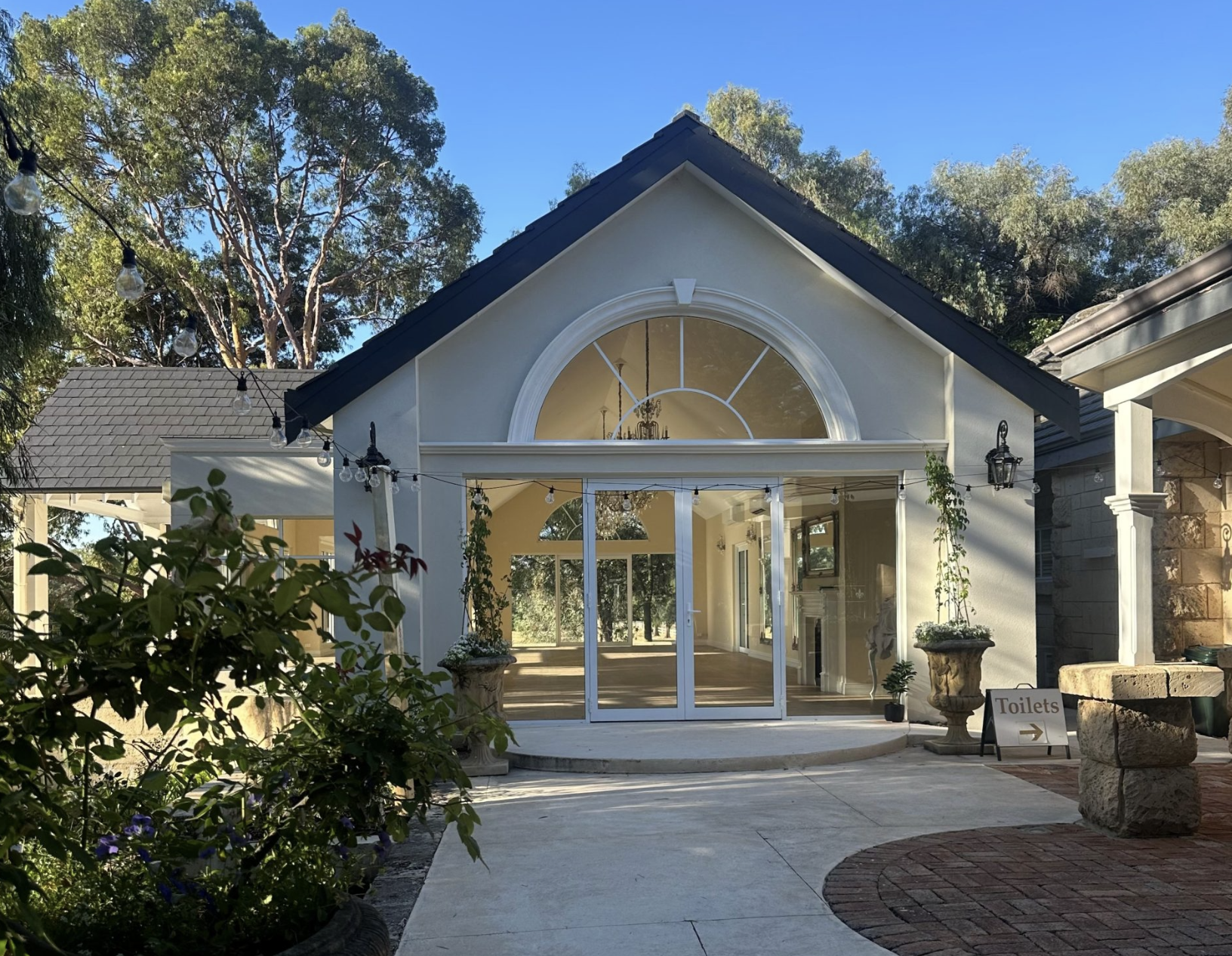 A house with a large arched window and glass doors, surrounded by greenery and outdoor string lights, with a sign indicating the location of toilets to the right.