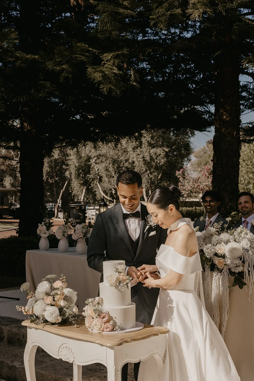 A couple in wedding attire cutting a wedding cake outdoors during daytime, with friends and floral arrangements in the background.