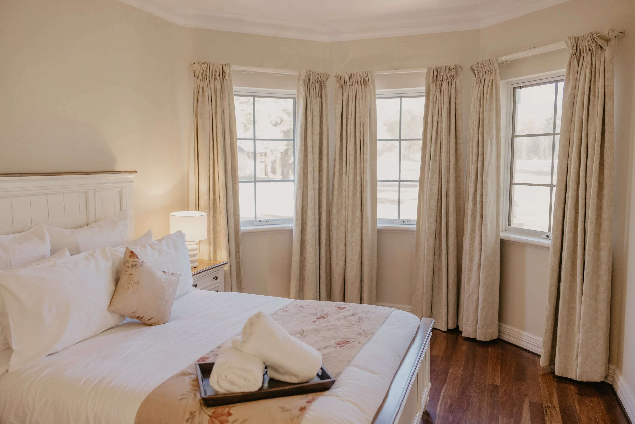 A bedroom with a bed covered in white linens and pillows, a side table with a lamp, and three large windows with light-colored patterned curtains, allowing natural light into the room.
