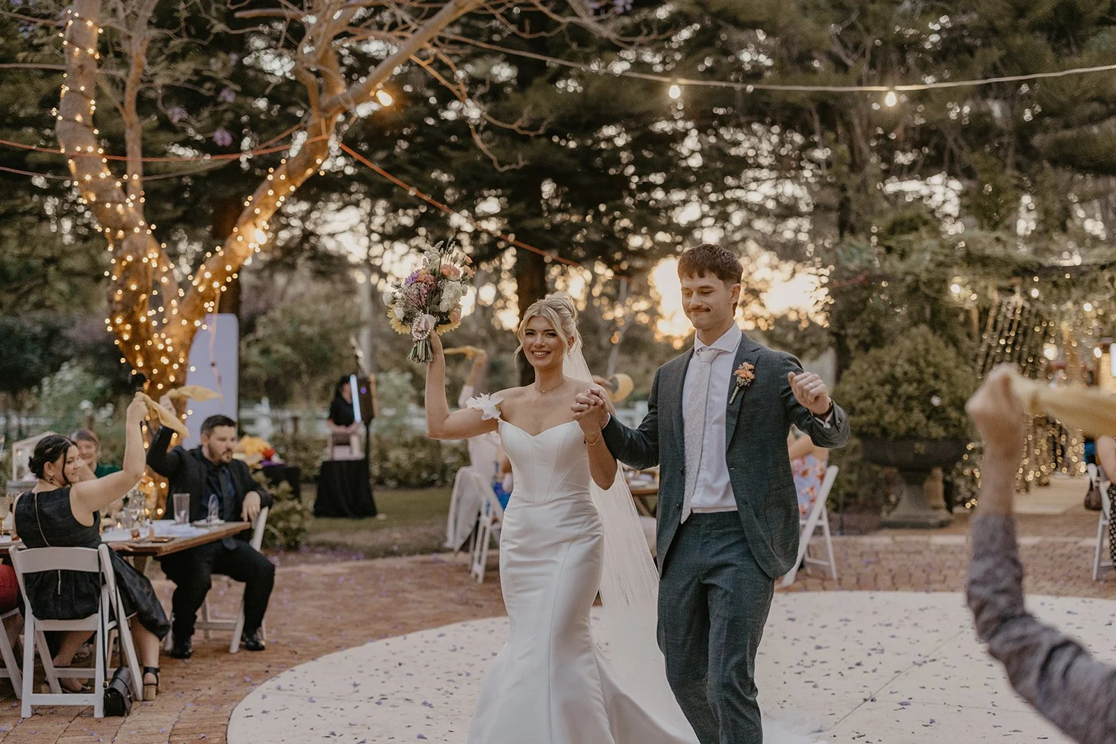 A bride and groom dance outdoors at their wedding reception during sunset, with guests seated at decorated tables and string lights hanging in trees.