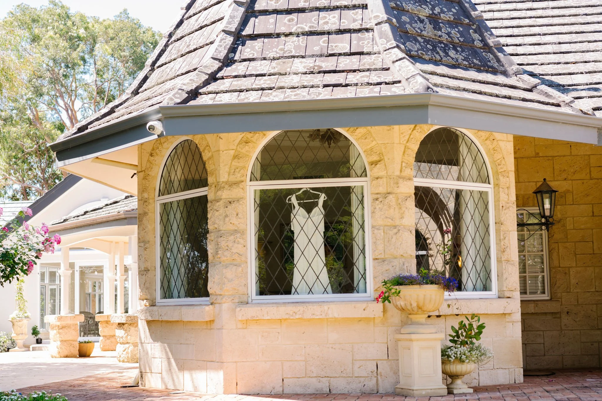 Front view of a house with arched windows, a stone facade, and a steep sloped roof. There are potted plants with flowers near the windows and a lantern-style exterior light on the wall.