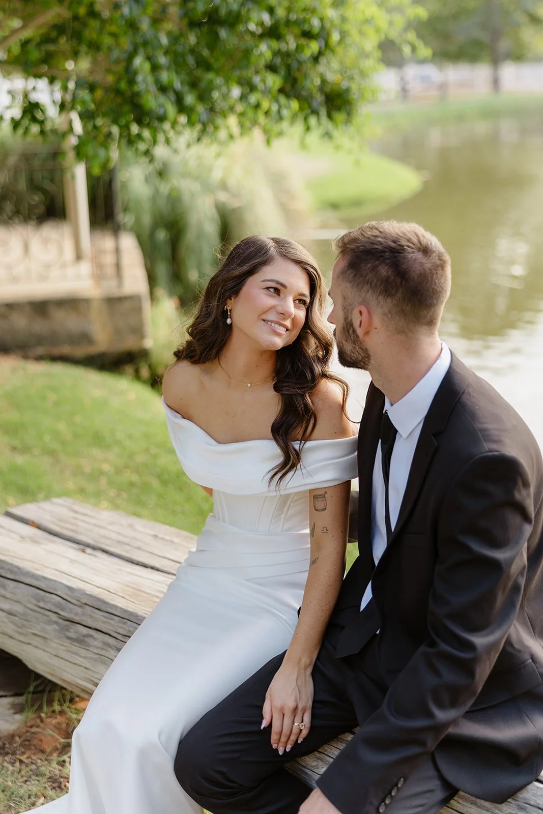 A bride and groom sitting on a wooden bench near a pond, gazing at each other with smiles, surrounded by greenery.