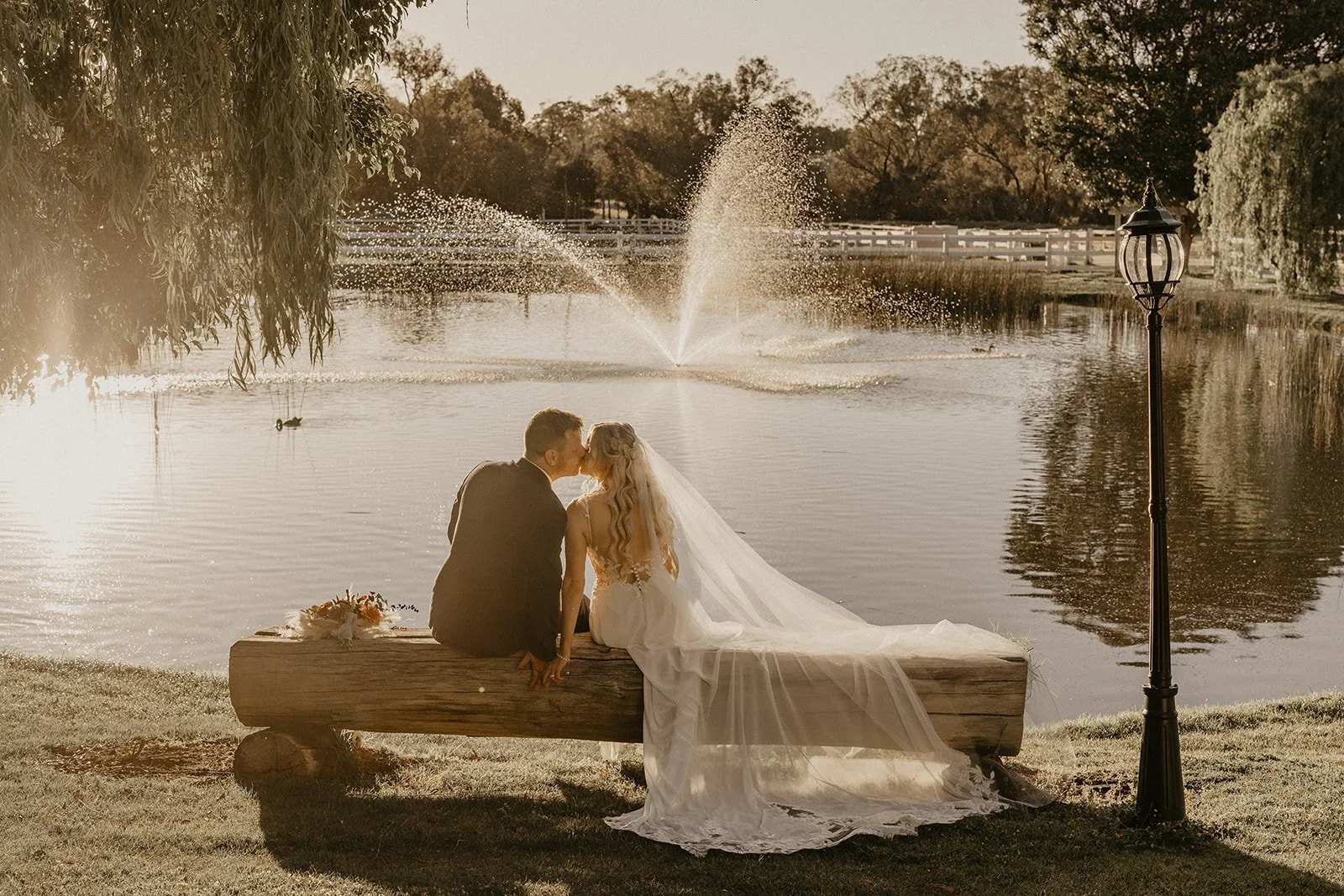 A bride and groom sitting on a wooden log by a pond, kissing with a fountain spraying water in the background, during sunset.