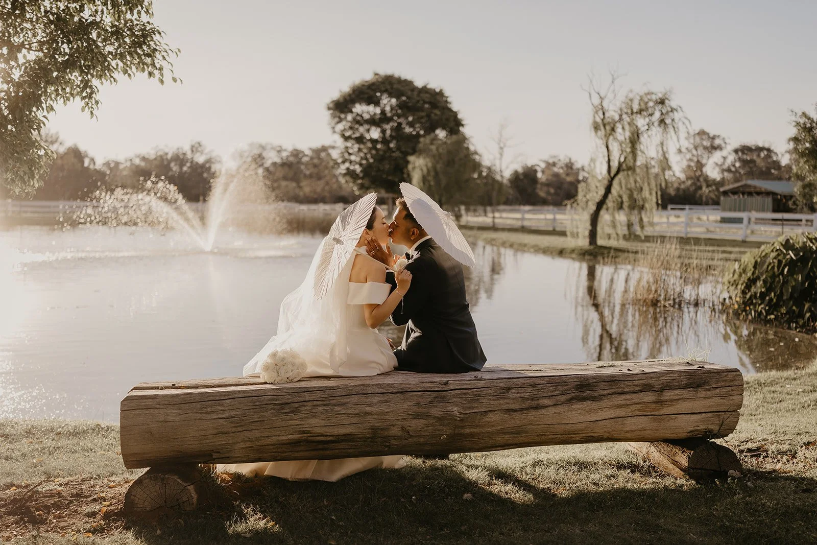 A bride and groom sitting on a wooden log bench by a pond, sharing a kiss and holding white parasols, with a fountain and trees in the background during sunset.