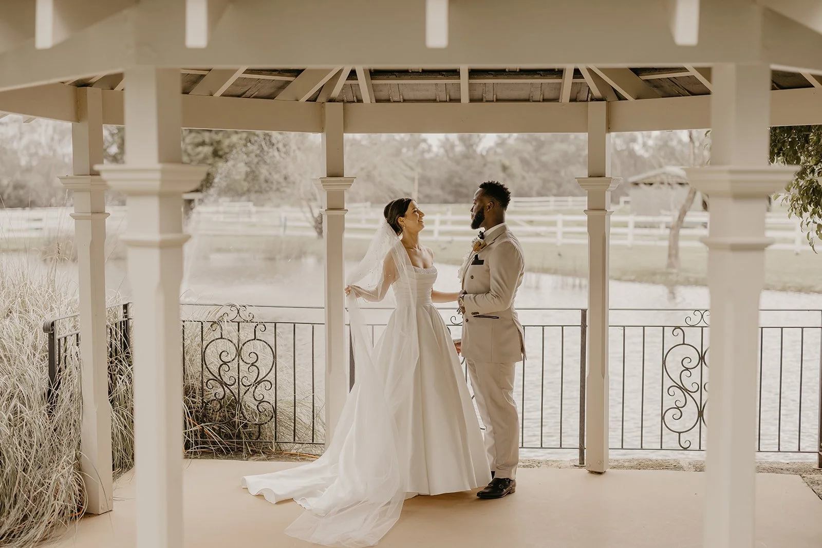 Bride and groom standing face-to-face under a gazebo by a pond, exchanging vows or sharing a moment during their wedding.