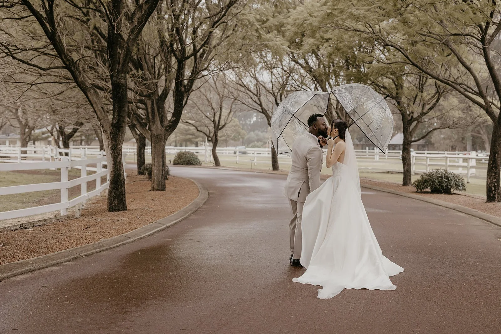 A bride and groom standing close together under umbrellas on a park road with trees and fencing in the background.