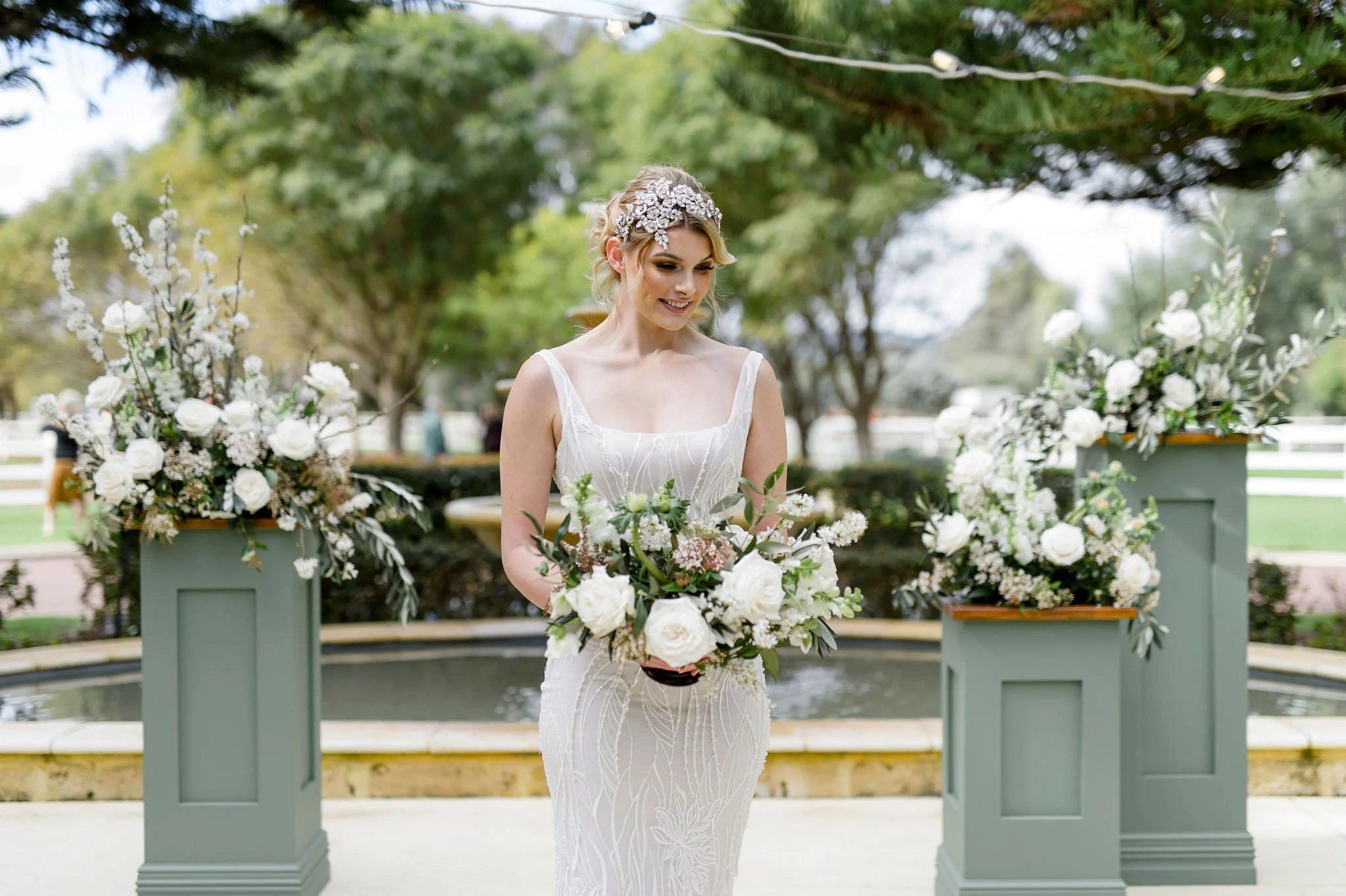 A bride holding a bouquet of white and pink flowers, wearing a white wedding dress with intricate patterns, standing outdoors surrounded by white floral arrangements and greenery.
