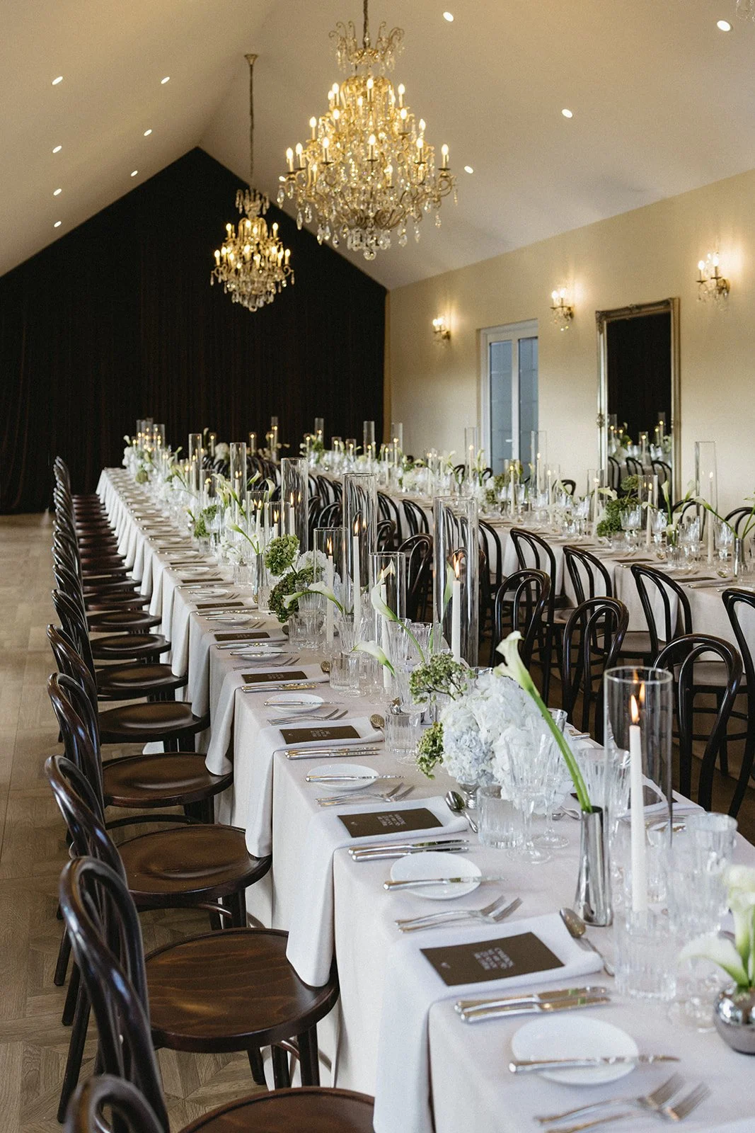 Elegant banquet table setting with white tablecloths, floral arrangements, tall glass candle holders, and cutlery in a decorated event hall with chandeliers.
