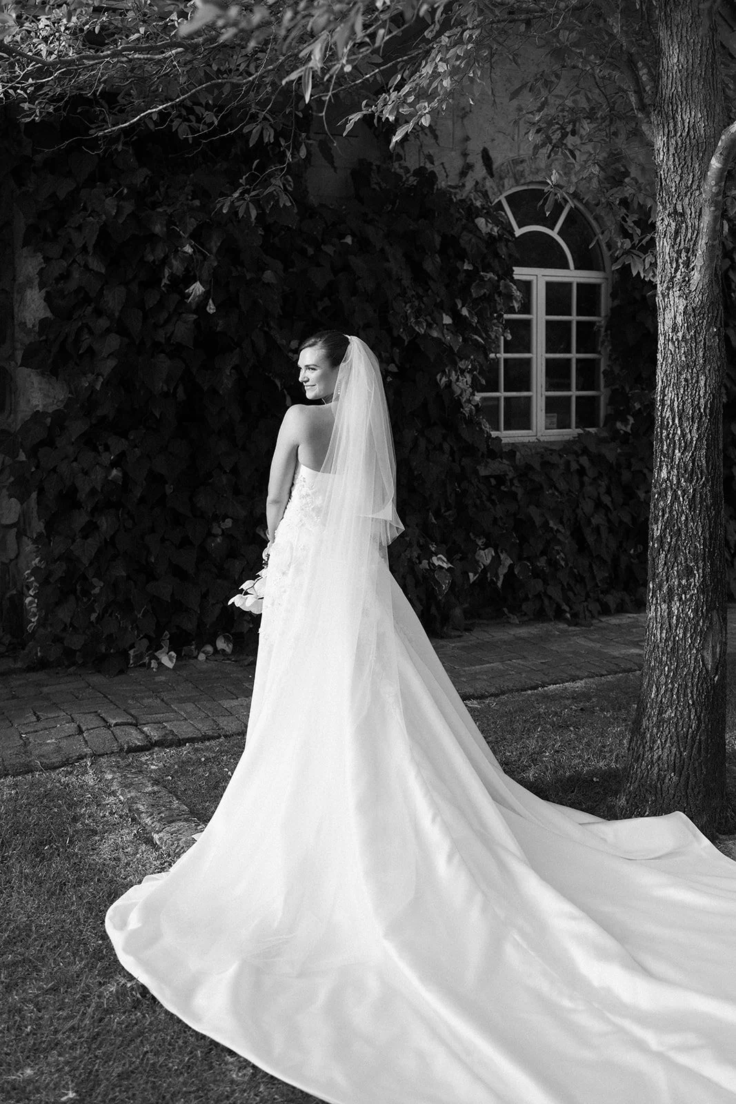 A bride in a wedding dress with a veil, standing outdoors at night near a tree and a window, smiling and looking over her shoulder.