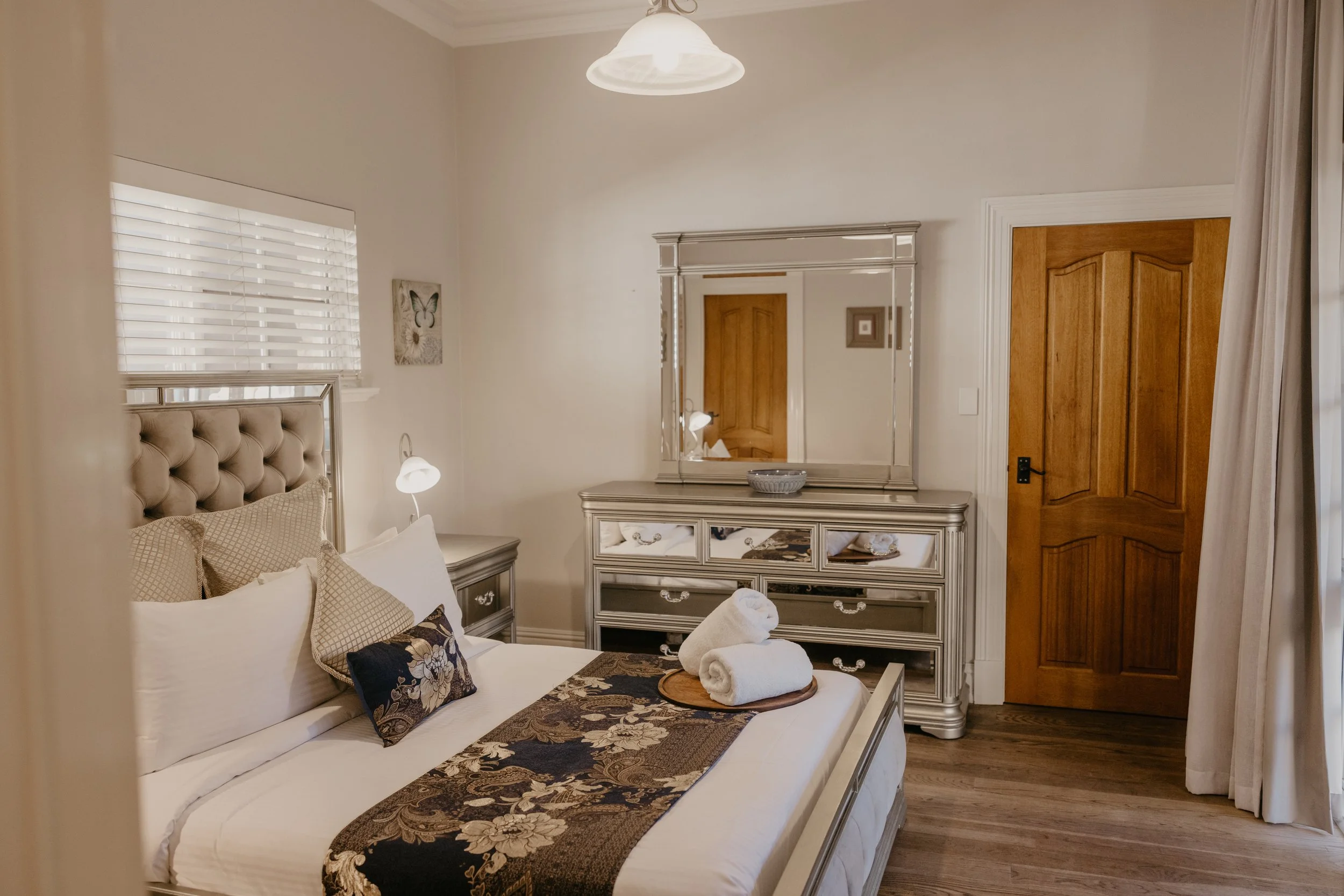 A cozy bedroom with a beige upholstered headboard, white bedding, patterned pillows, and a floral bed runner. There are rolled towels on a small round table at the foot of the bed. A silver dresser with a mirror, a small bowl, and a decorative drawer