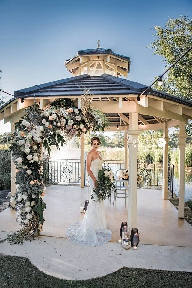 A bride in a white wedding dress holding a bouquet stands under a gazebo decorated with white and blush flowers, set outdoors on a sunny day.