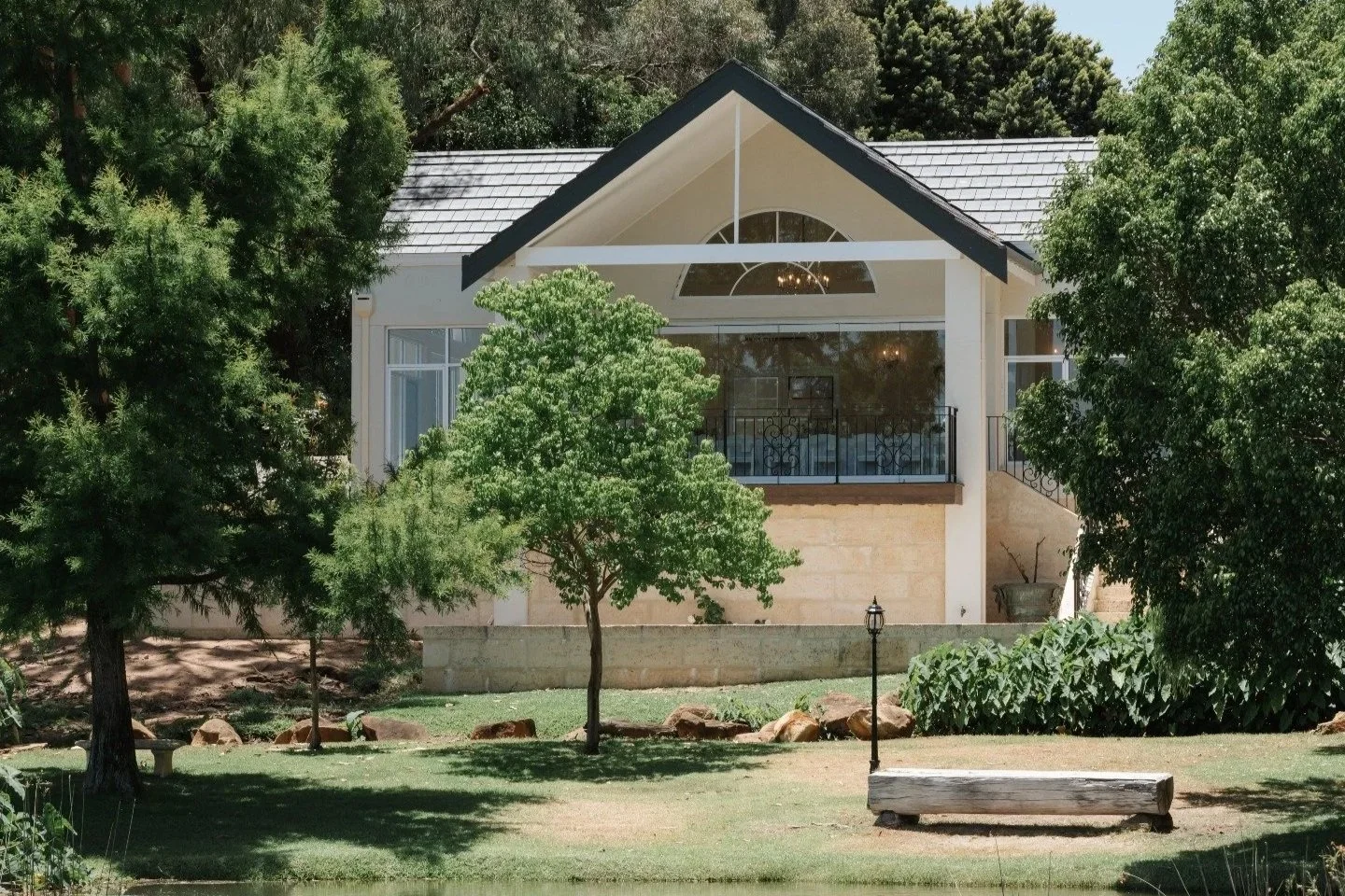 A house with a large front window and a small front porch, surrounded by trees and a grassy yard with rocks and a wooden bench.