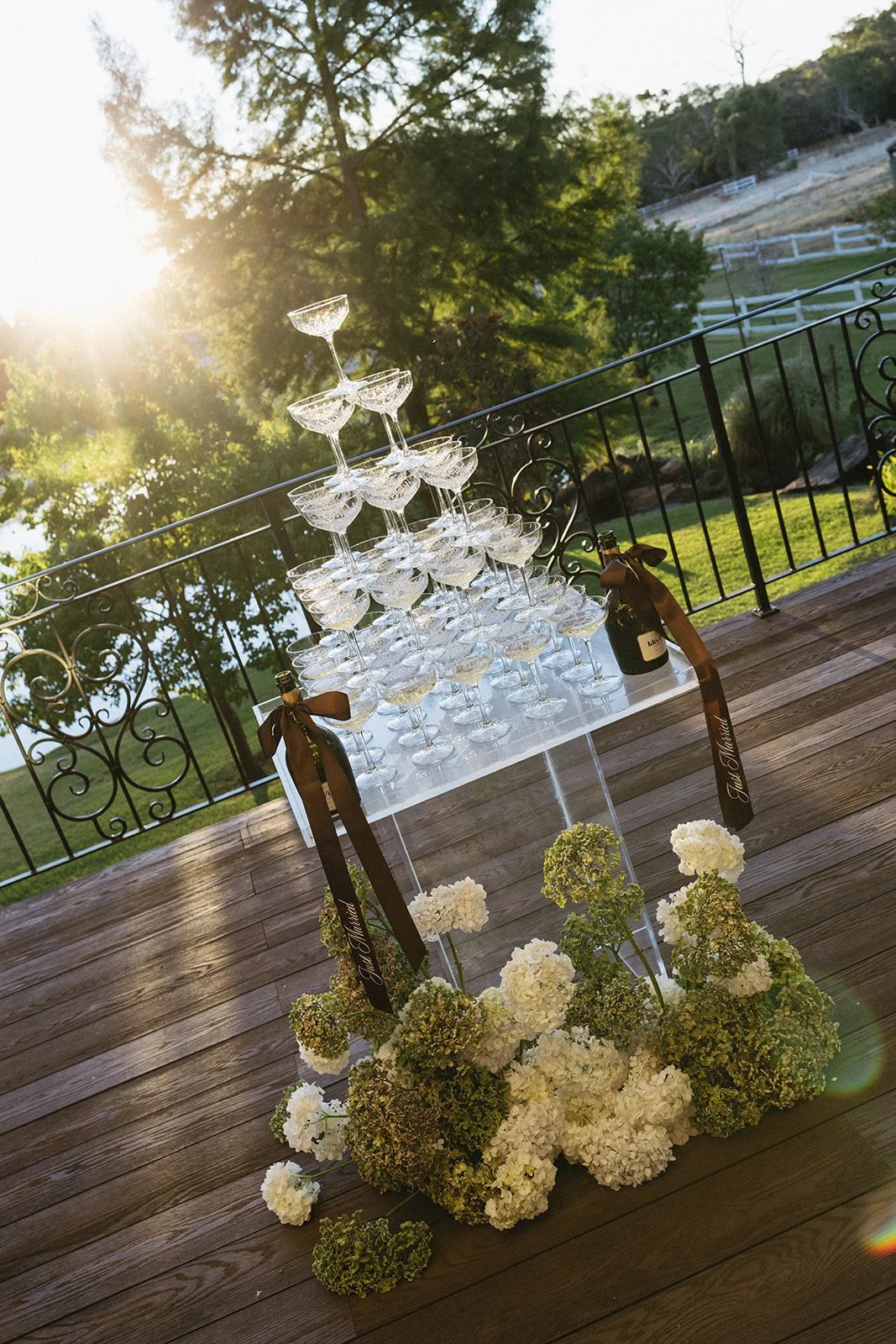 A pyramid of empty champagne glasses on a white table, decorated with white hydrangeas, two bottles of champagne with ribbons, set on a wooden deck with a scenic outdoor background of trees and a white fence at sunset.