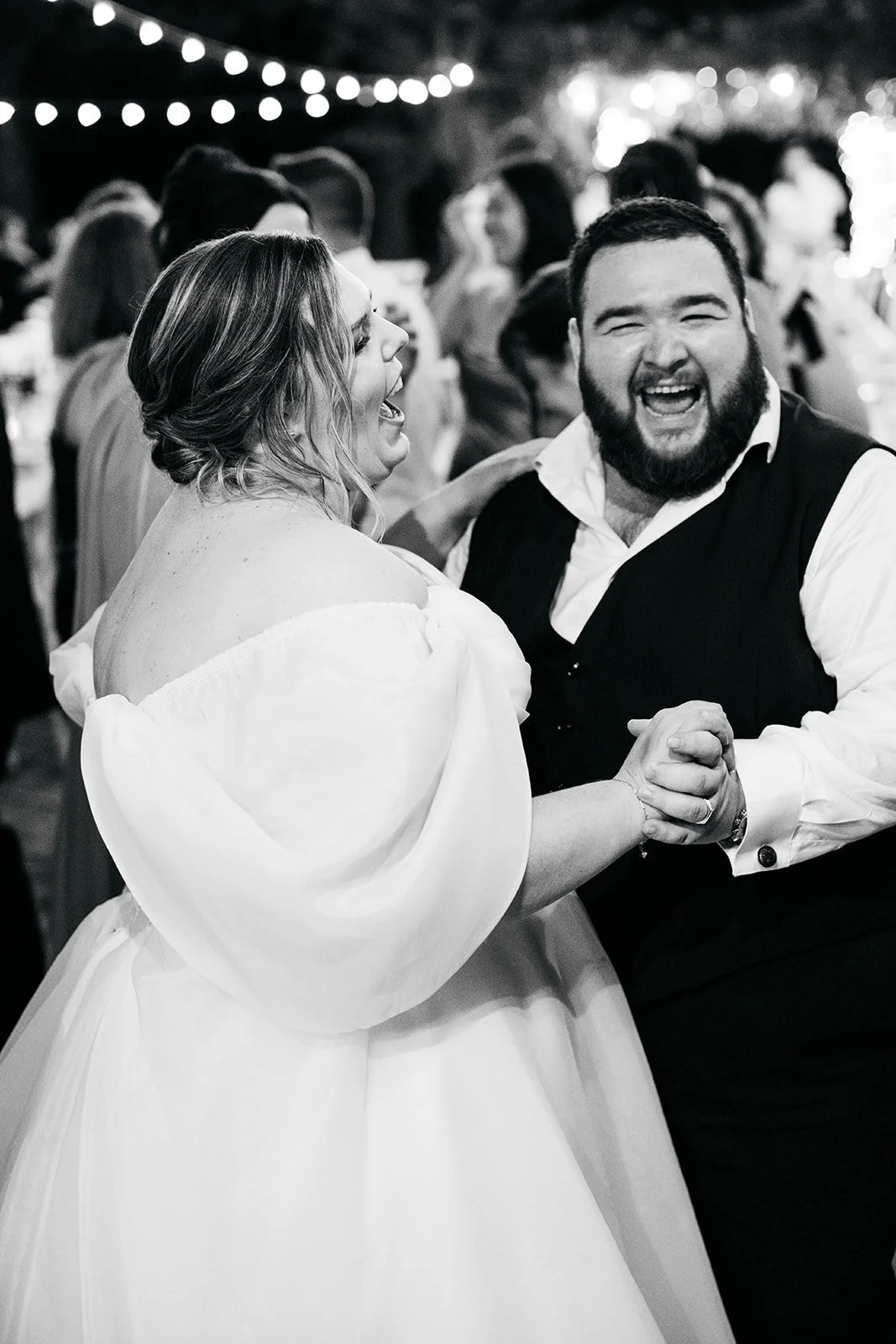 A couple dancing at their wedding reception, smiling and holding hands, with string lights hanging overhead and guests in the background.