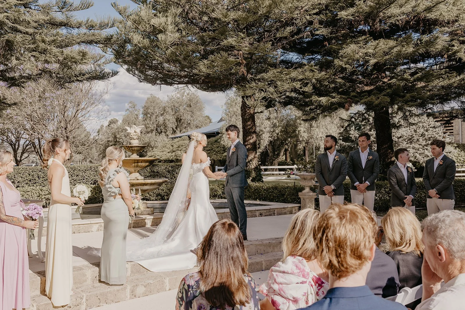 A wedding ceremony outdoors with the bride and groom holding hands in front of guests. The bride is wearing a white wedding dress with a long veil, and the groom is in a suit. Bridesmaids and groomsmen stand on either side, with the bridesmaids holdi