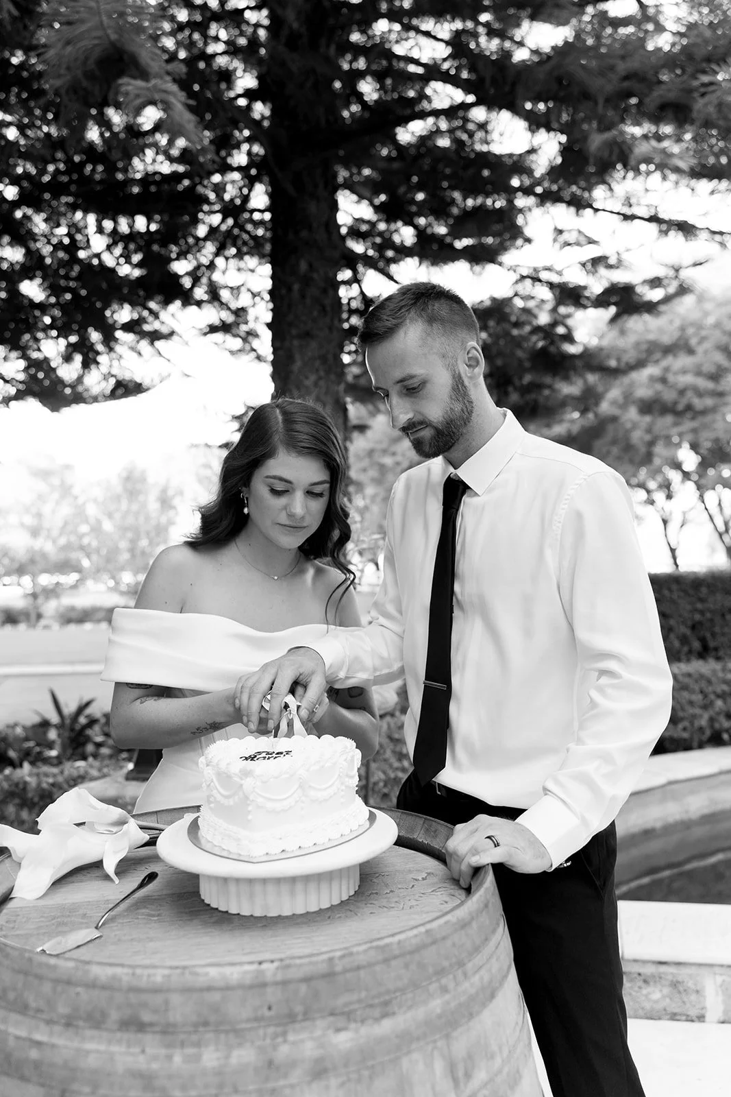 A couple at a wedding cutting a cake outdoors under trees, black and white photograph.