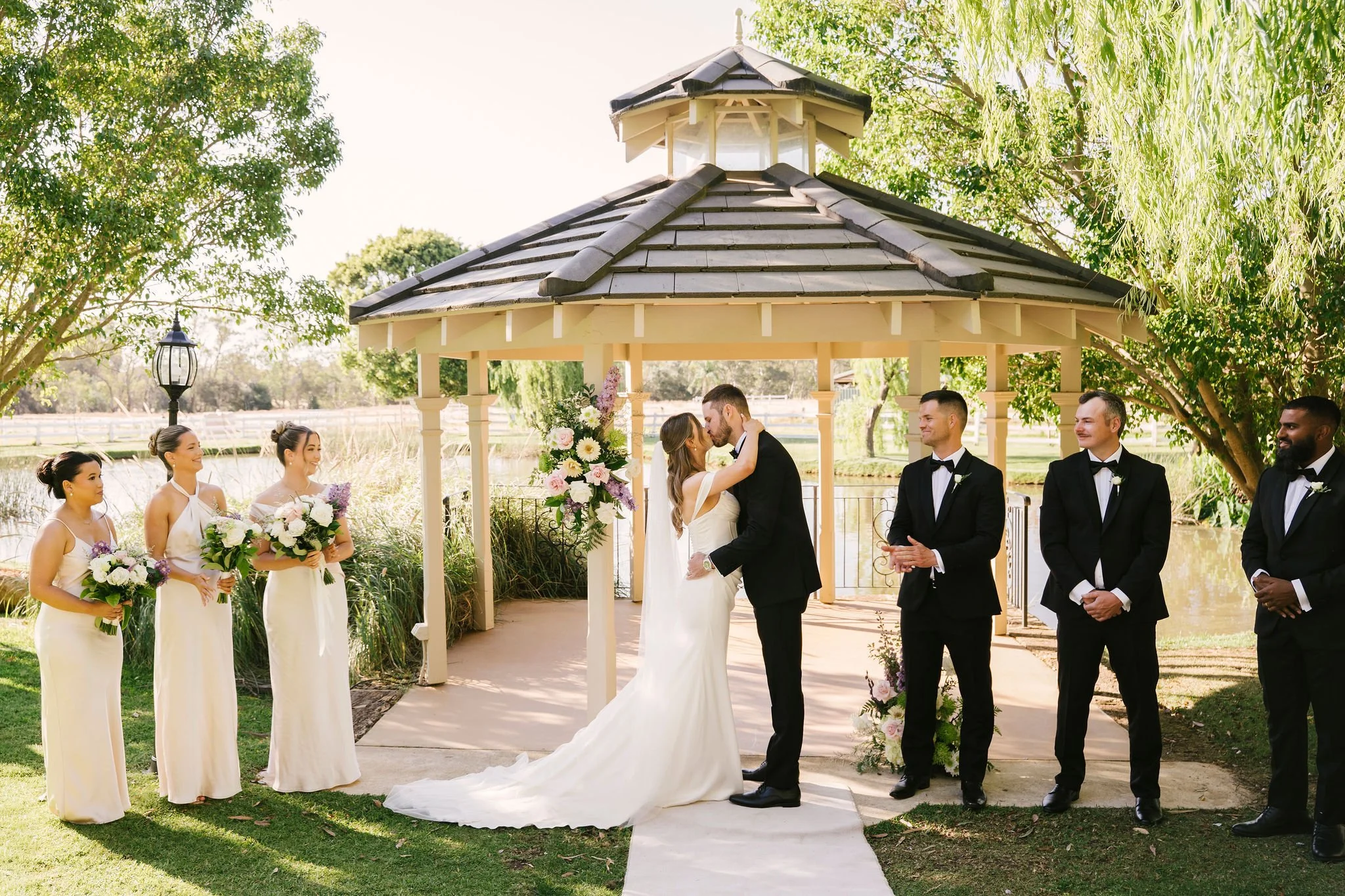 A wedding ceremony taking place outdoors under a gazebo with a bride and groom kissing, surrounded by bridesmaids and groomsmen.