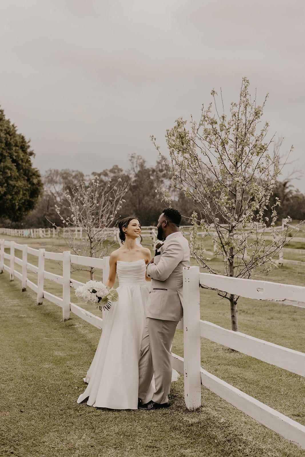 A bride in a white wedding dress and a groom in a beige suit sharing a romantic moment outdoors near a white wooden fence and blossoming trees.