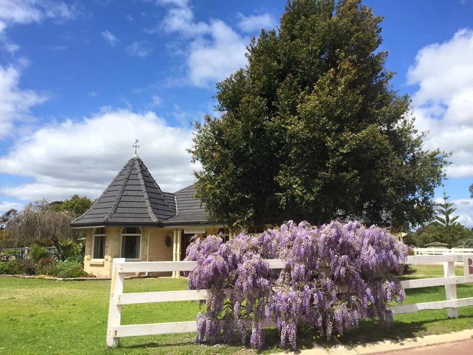 A small building with a pointed roof and a cross on top, surrounded by green grass, a large tree, and a bush with purple flowers, under a partly cloudy blue sky.