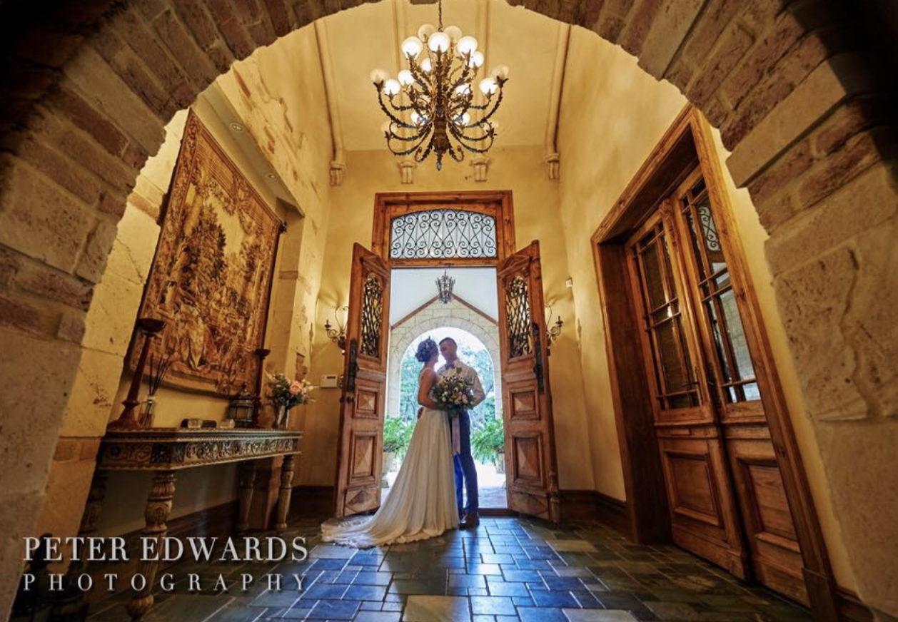 Couple in wedding attire standing in a decorative entryway with wooden doors, ornate chandelier, and vintage furnishings.