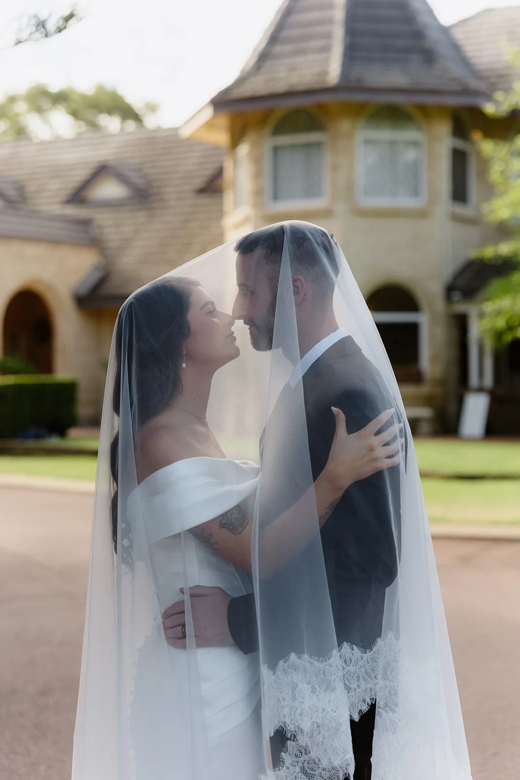 A bride and groom, wrapped in a bridal veil, embrace and look into each other's eyes in front of a large, ornate house with a turret, lush greenery, and cloudy sky.