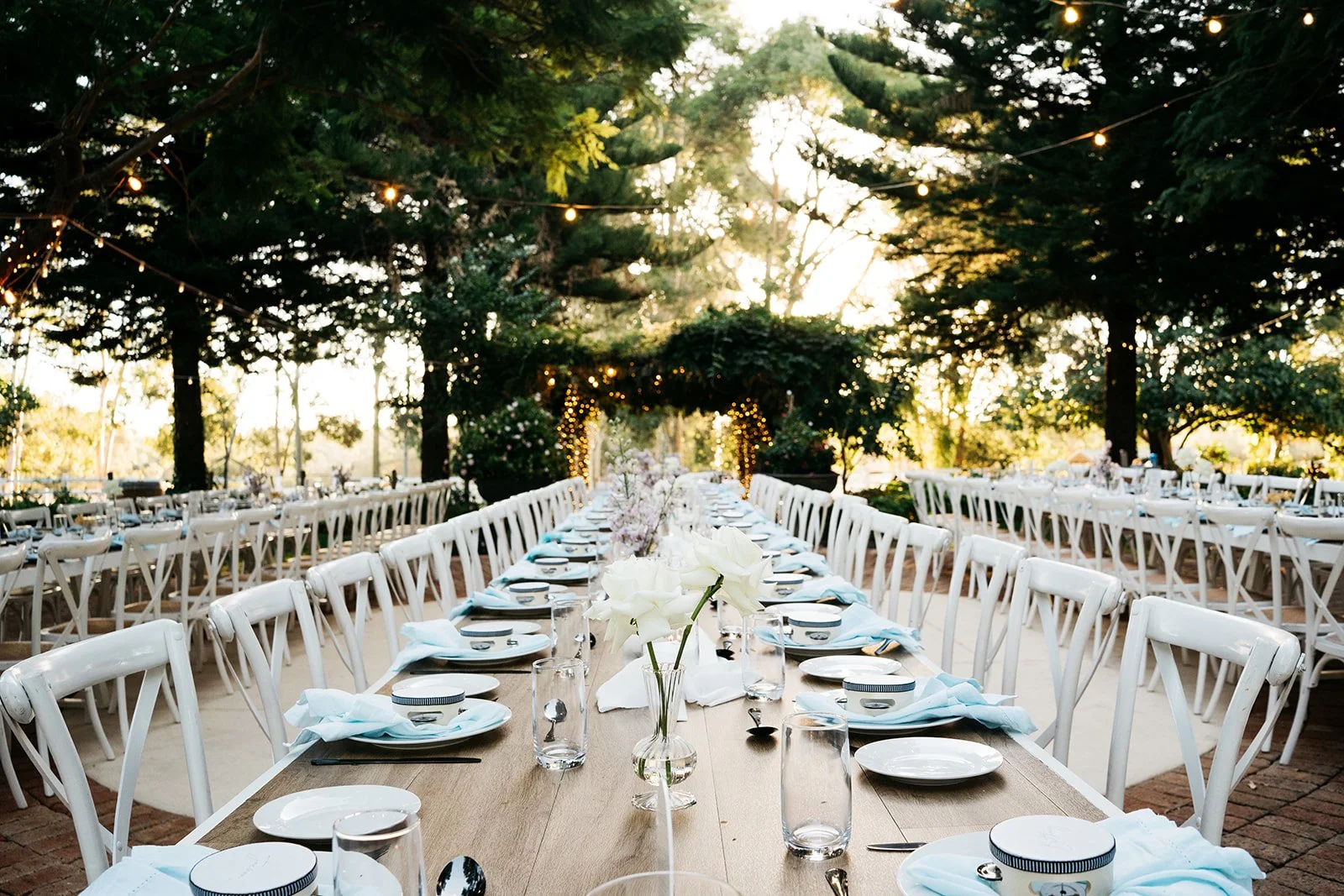 Outdoor dining setup with long wooden tables, white chairs, and floral centerpieces under trees with string lights, during sunset.