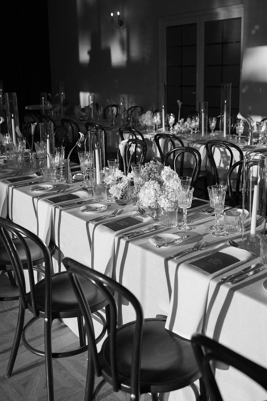 A black and white photograph of an elegantly set banquet table with flowers, glassware, and tableware in a dimly lit room.