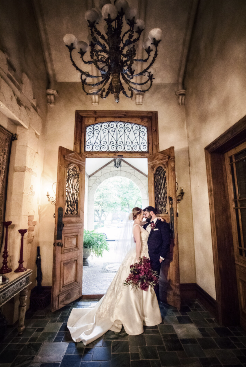 A bride and groom standing close together in a doorway, holding a bouquet of flowers, with open wooden doors and a chandelier overhead.