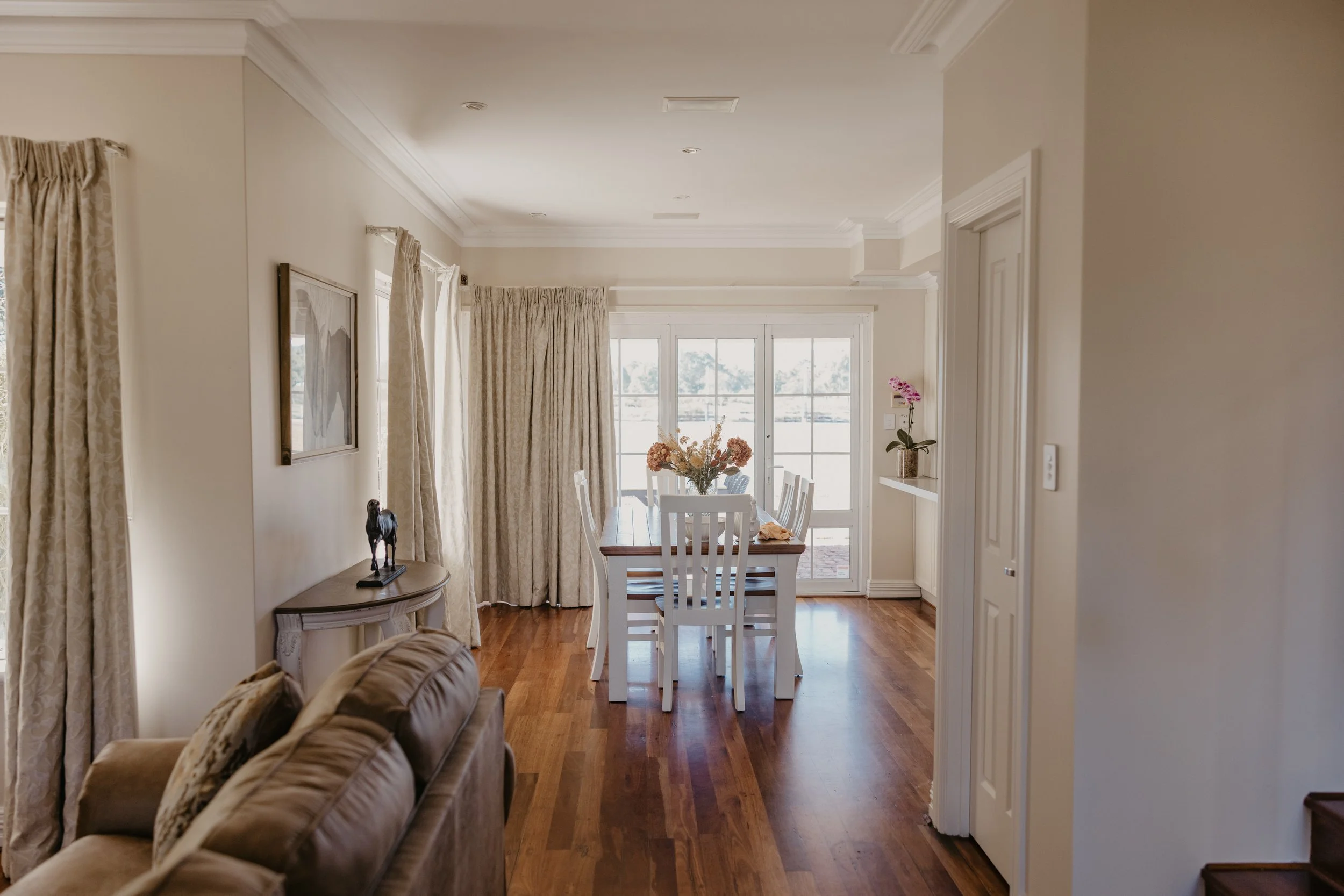 A dining area with a table and chairs in front of large windows and glass doors, decorated with flowers, with cream curtains and crown molding on the ceiling.