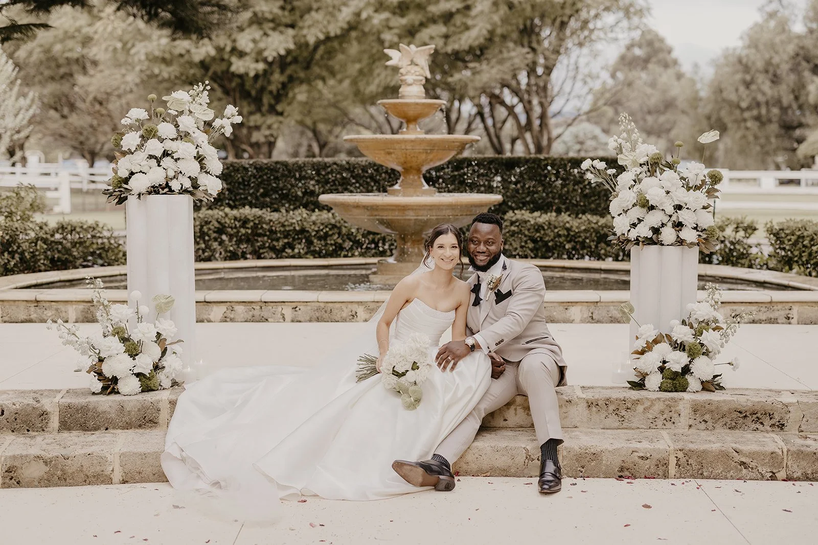 A bride and groom sitting together on stone steps in front of a large fountain with tiered shelves, outdoors with trees and a white fence in the background. The bride is holding a bouquet of white flowers and wearing a strapless white wedding gown. T